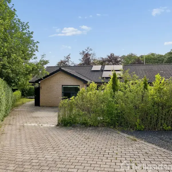 En gul murstensvilla med solpaneler står stille, omgivet af en plan indkørsel og grønne buske. Foran er en flisebelagt sti med tætklippede hække på begge sider under en blå himmel.
