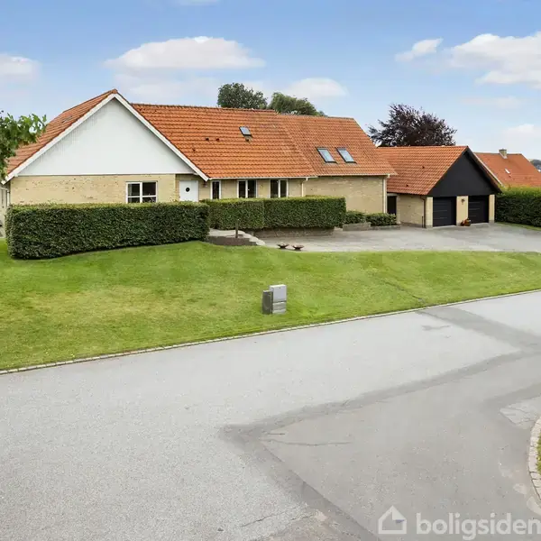 Hus med rødt tegltag og gule mursten står på en velholdt grøn plæne. Indkørsel leder til en dobbeltgarage. Omgivet af hække og træer under en blå himmel.