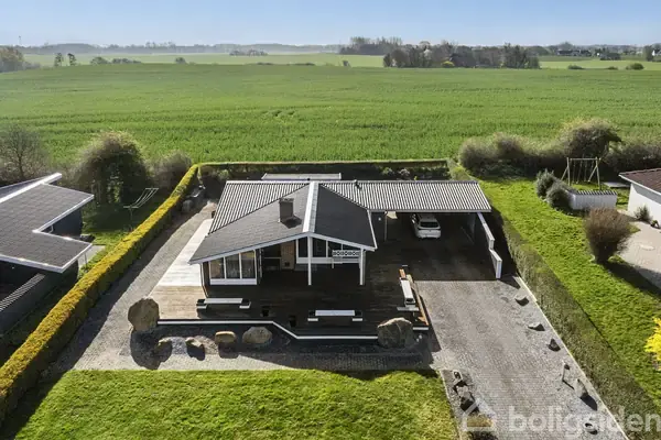 Huset står stille med sort tag og hvid facade, omgivet af en stenbelagt gårdsplads og grøn have. Baggrunden består af store, åbne marker.