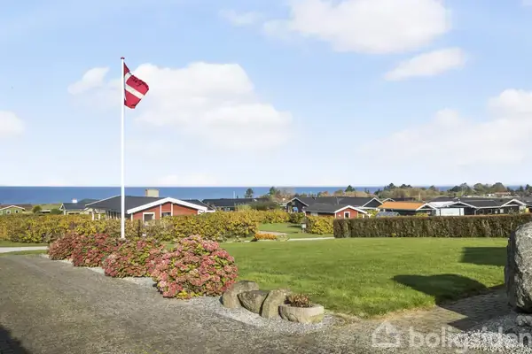 Flagstang med dansk flag vajer let i vinden på en velplejet have omkranset af buske og græsplæne. I baggrunden ses huse og havet under en klar, blå himmel.