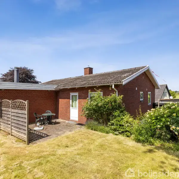 Rødt murstenshus, står på solrig græsplæne, med terrasse og havemøbler. Omkranset af buske og træer. Klar blå himmel i baggrunden.