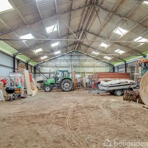 En traktor står parkeret i en stor lade med højt tag og åbne bjælker. Omkring traktoren er der værktøj, halmballer og forskelligt landbrugsudstyr på den jordede gulvflade.