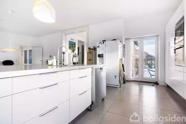 Et moderne køkken har en stor køkkenø med vask og stilrene skabe. I baggrunden ses en køleskab, opvaskemaskine og terrassedøre, der giver lysindfald.