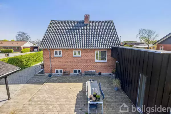 Et rødt murstenshus med sort teglstenstag står i en gårdhave med fliser. En udendørs spisebord er placeret i midten, omgivet af enkelte planter i krukker.