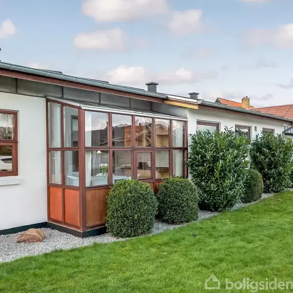 En hvid bungalow med flere store vinduer har en lukket veranda. Den står på en velplejet græsplæne med små buske langs væggen. En mælkekande er placeret ved siden af huset.
