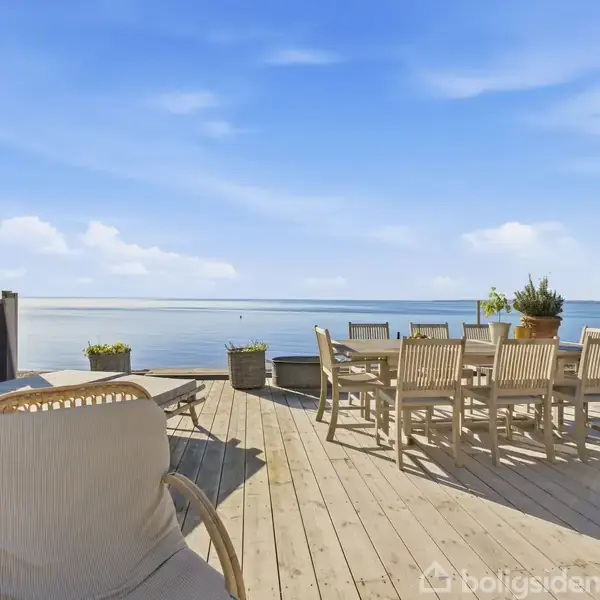 Træterrasse med havemøbler, hviler ved havet. Et bord og stole står under den blå himmel. Planter i potter skaber en hyggelig atmosfære.