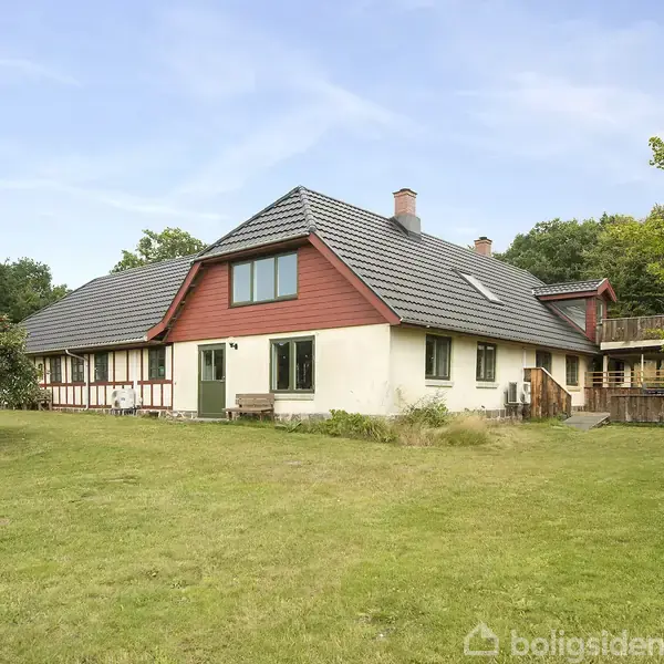 Et stort hus med rødt tag og hvide vægge står i en grøn have. Træer omgiver ejendommen, og himlen er blå.