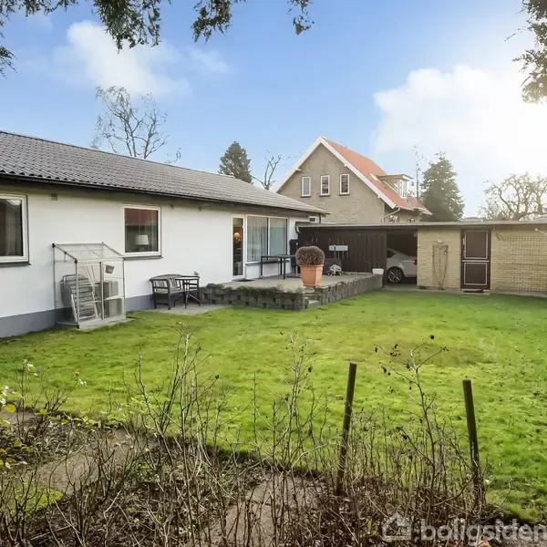 En hvid bungalow med fladt tag står i en have med græsplæne og buske. Der er en terrasse med havemøbler, et redskabsskur og en carport til højre.