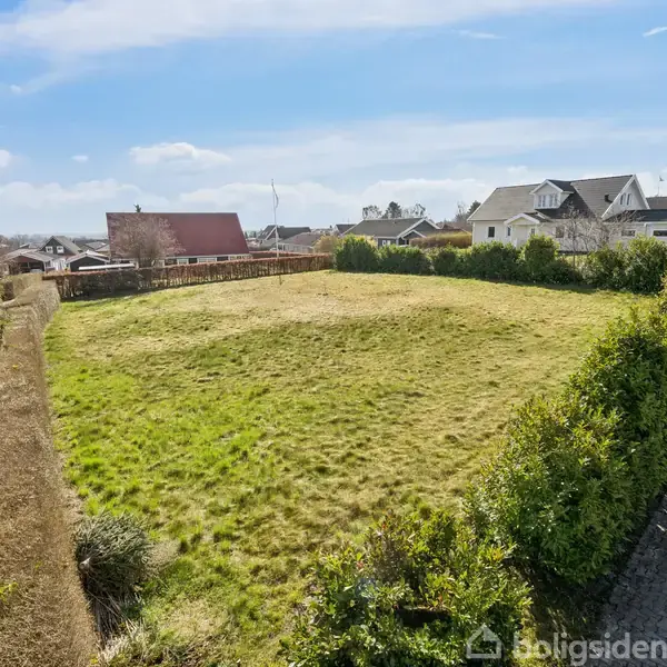 Græsplæne ligger stille omgivet af tætklippede hække. I baggrunden ses flere huse i et villakvarter under en klar blå himmel med enkelte skyer.