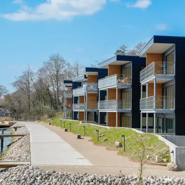 Lejlighedskompleks med moderne altaner står langs en gangsti. Stien strækker sig ved en vandkant, flankeret af grus og græs, mens træer og bygninger ses i baggrunden under blå himmel.