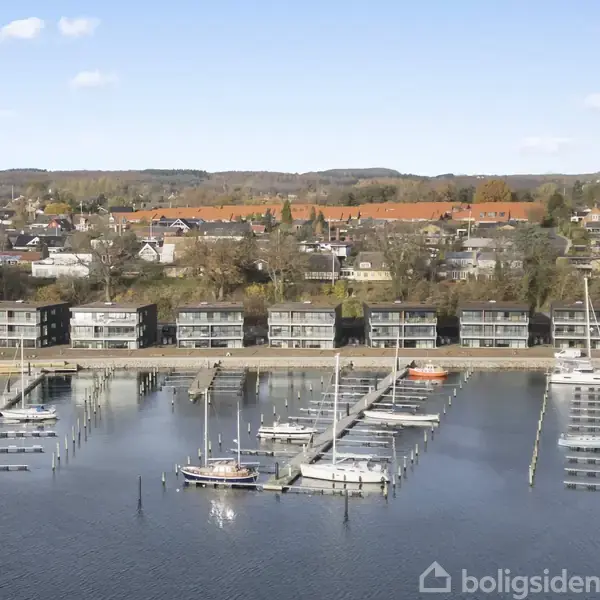 Lejlighedskomplekser placeret langs en havn ved vandet, hvor både ligger til kajs. I baggrunden ses en by med huse spredt blandt træer og et bakket landskab.