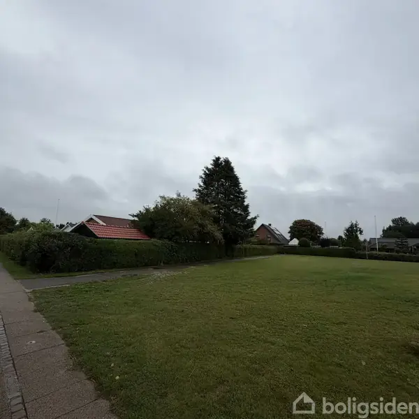 En åben græsplæne foran huse med røde tage og omkranset af grønne buske og træer, under en overskyet himmel.