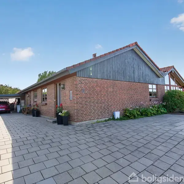Et rødt murstenshus med gråt tag står på en flisebelagt indkørsel. Biler er parkeret under en carport til venstre, omgivet af grønne hække og buske.