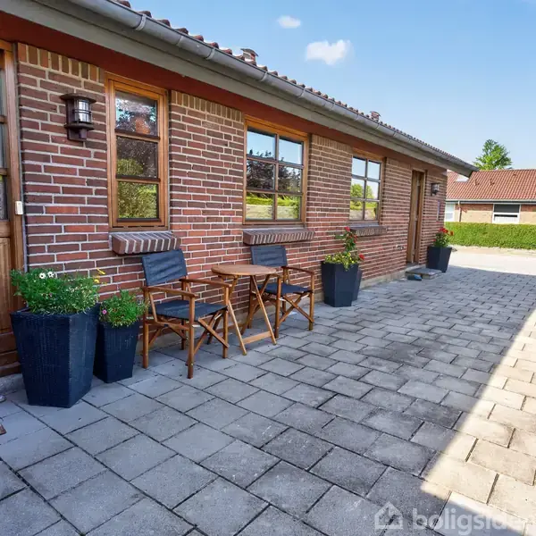 To træstole og et bord står på en flisebelagt terrasse foran et rødt murstenshus. Store krukker med planter flankerer møblerne; husets vinduer reflekterer sollyset.