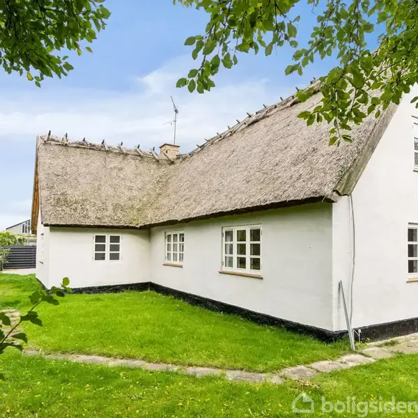 En traditionel, hvidmalet stråtækt hus står omgivet af en grøn have og træer. Huset har store vinduer og en firkantet form, beliggende i et roligt boligområde under en blå himmel.