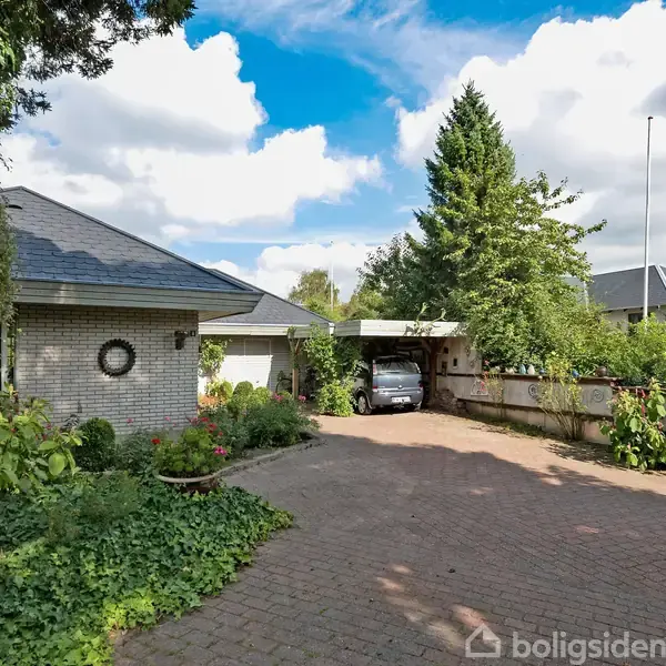 Hus med gråt murstensvæg og skråt tag. Bil parkerer under en carport omgivet af frodig have med blomsterbede. Klar himmel med få skyer.