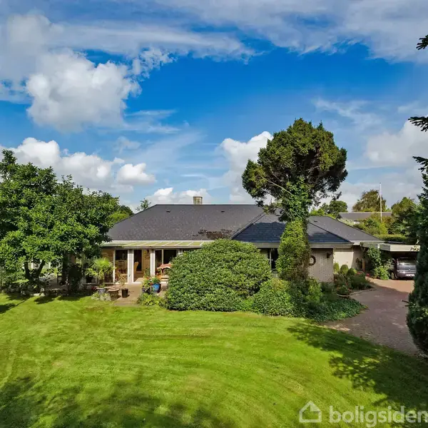 Et moderne hus med fladt tag står i en frodig have med grønne plæner og træer. Blå himmel og skyer danner en klar baggrund.