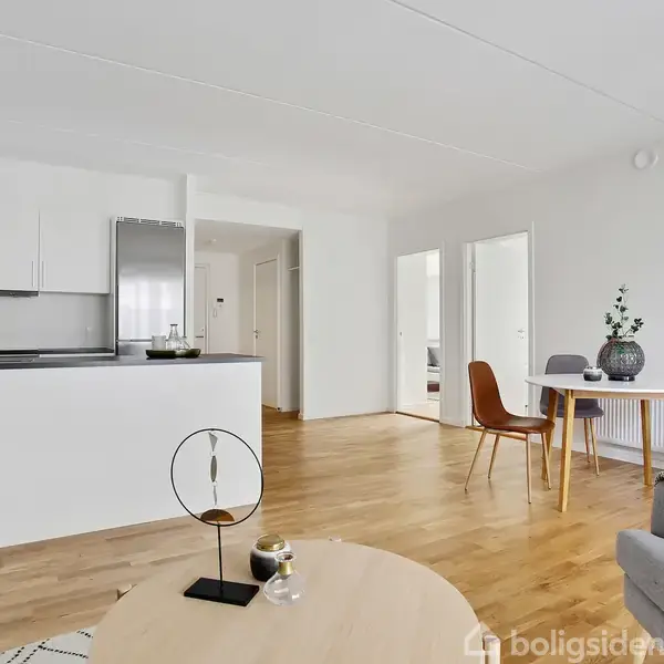 Stue med sofabord, vase og to sofaer, handling: indrettet minimalistisk. Kontekst: hvide vægge, åbent rum med køkken, spisebord med stole i baggrunden, trægulve.