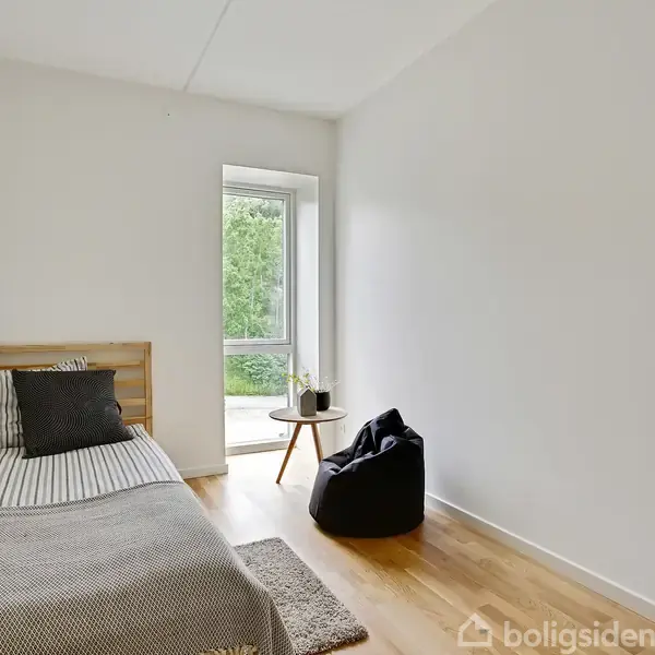 Seng står ved væggen i et minimalistisk værelse. En stor pude ligger ved siden af et lille bord. Et vindue viser grønne træer udenfor. Ingen tekst til stede.