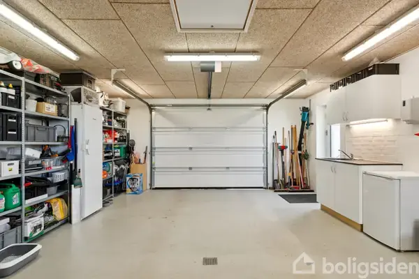 Garage med opbevaringshylder fyldt med værktøj og materialer; en stor port lukket. Væggen til højre har vask, skabe og rengøringsredskaber stående ved siden af.