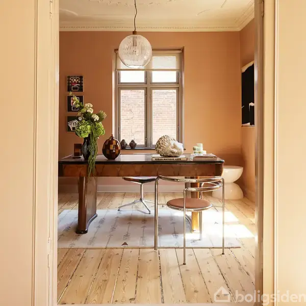 Et skrifbord står i et lyst rum med træplankegulv. På bordet er vaser og skulpturer. En vindueskarm med lys strømmer ind fra et stort vindue. Bøgerne inkluderer "HOLLAND INTERIOR GOTH".