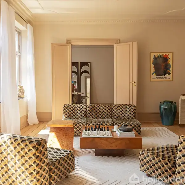 En sofa med mønstret stof står i et værelse med lyse vægge. Foran sofaen er et sofabord med et skakbræt. Til højre hænger en farverig plakat.