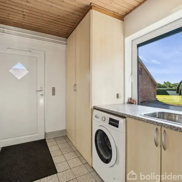 Vaskemaskine står under køkkenbord ved siden af vask. Værelse med hvide skabe, hvid dør, flisegulv, og stort vindue med udsigt til have og blå himmel.