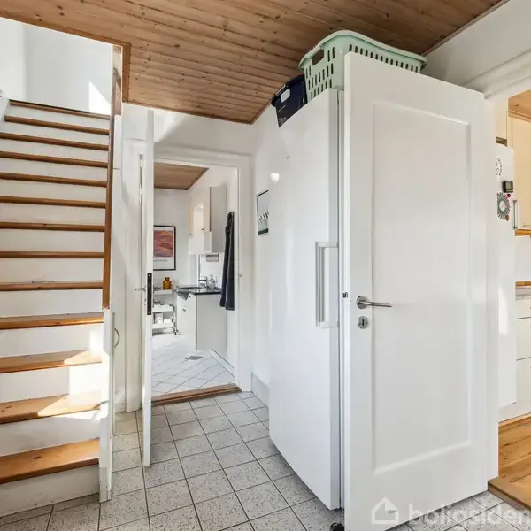 Trappe fører opad ved siden af et køkken med hvide skabe. En kurv ligger ovenpå et højskab. Flise- og trægulve dækker gulvet i en lys korridor.