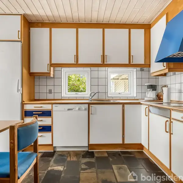 Køkken med hvide skabe, blå detaljer og ovn, arrangeret langs en væg med to små vinduer. Flisebelagt væg og bordplader, spisebord med blå stole i forgrunden.