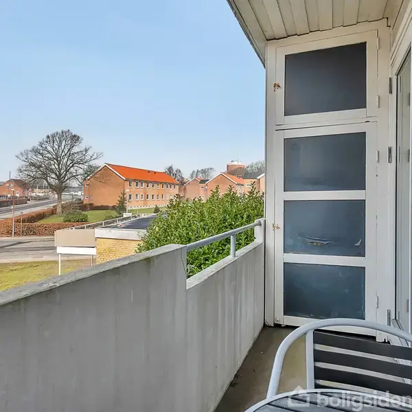 Balkon med et lille bord og stol, omgivet af et hvidt rækværk. Udsigt over en vej med rødtagsbygninger og et træ i et boligområde.