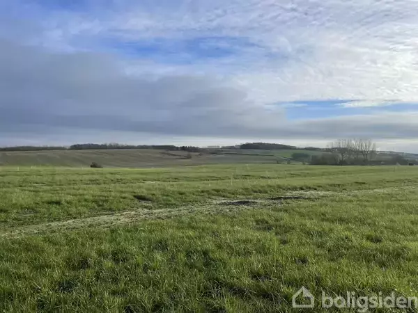 Grøn mark strækker sig over et bakket landskab under en delvist skyet himmel, med spredte træer og fjerne bakker i horisonten.