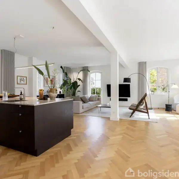 En stue med et åbent køkken, hvor køkkenøen står centralt. Der er moderne møbler, store vinduer, planter og kunst på væggene. Tekst på billede inkluderer “LOVE IS IN MY HOME.”