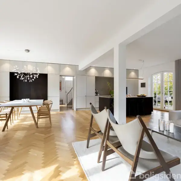 Et moderne og lyst stue-område med træmøbler og spisebord i forgrunden. Rummet har store vinduer, trægulve og en sofagruppe, der skaber en åben og indbydende atmosfære.