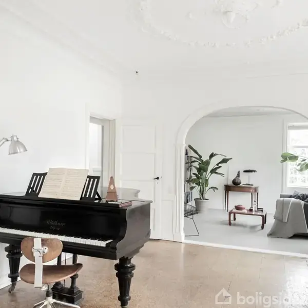 Klaver står stille overfor en buet døråbning. I baggrunden ses en lys stue med grønne planter og en grå sofa. På klaveret ligger noder.