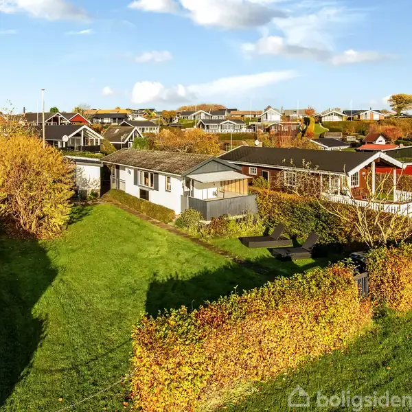 Et hvidt hus står stille omkranset af efterårsfarvede hække i en rækkehusbebyggelse. Græsplæner og flere huse ses i det solrige landskab.