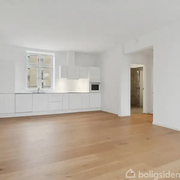 Et rummeligt, lyst køkken-alrum domineret af hvide skabe og en indbygget ovn. Rummet har trægulve og et stort vindue, der tillader naturligt lys at strømme ind.