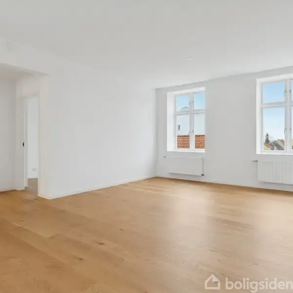 Et lyst værelse med trægulv har to vinduer, der lukker dagslys ind. Væggene er hvide, og der er en radiator under hvert vindue. I hjørnet ses en åbning til et andet rum.