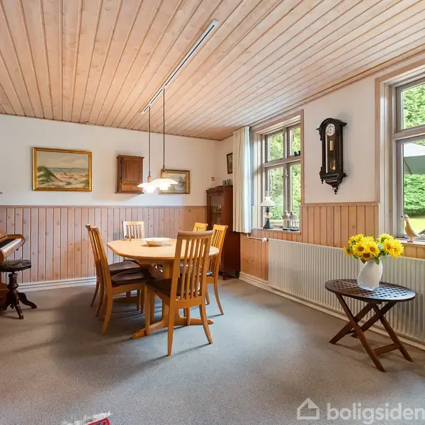 Et spisebord med træstole står i et lyst rum med træloft. Et klaver, malerier og en væg-ur dekorerer væggen. Store vinduer viser en grøn have. En vase med gule blomster står på et bord.