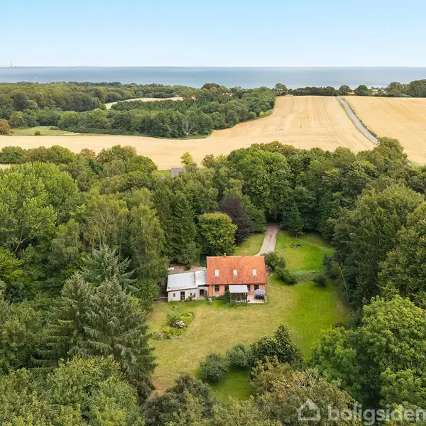 Et hus med rødt tag ligger omgivet af tæt skov. Det åbne græsareal foran huset skaber en lysning. Baggrunden består af dyrkede marker og en fjern udsigt over havet.