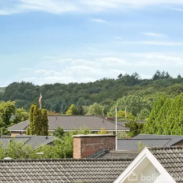 Et rødt murstenshus med skråt tag, omgivet af en grøn græsplæne. Huset har flere vinduer, og en terrasse ses i forgrunden. Baggrunden inkluderer en anden bygning.