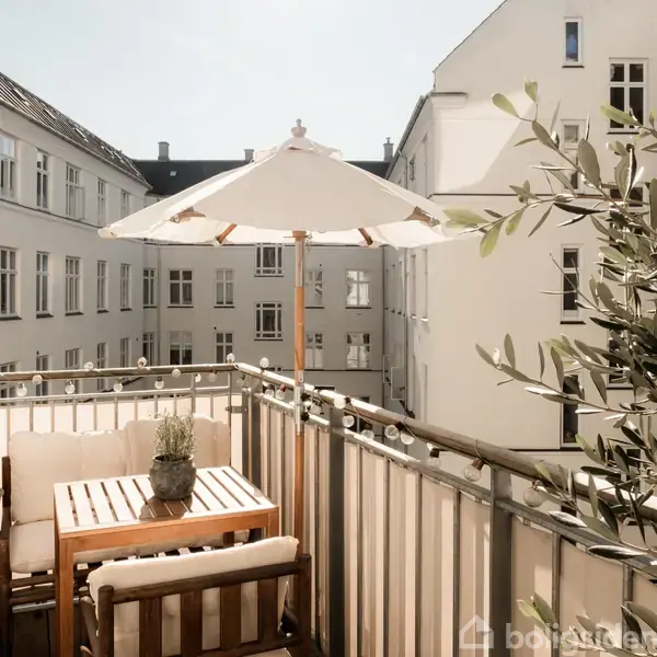 En terrasse med et bord og en plante i potte under en hvid parasol. Omgivet af bygninger med mange vinduer.