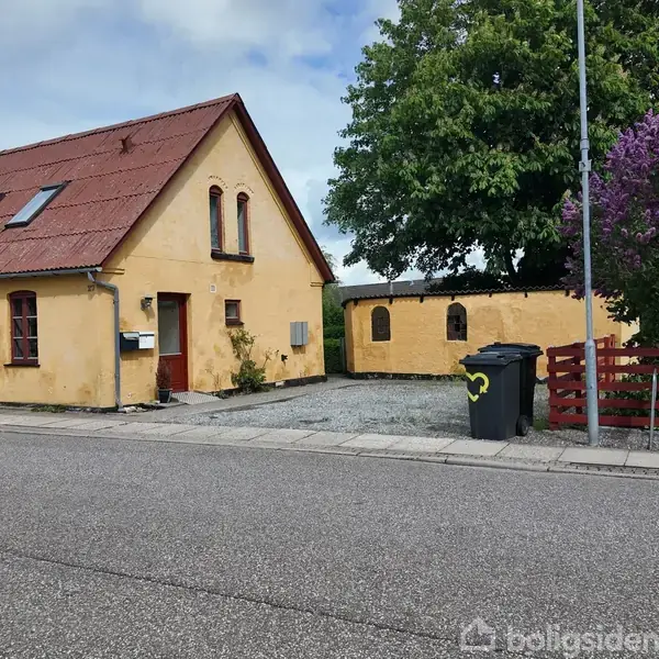 Et gulmalet hus står ved siden af en vej. En skraldespand med et gult hjertemotiv er placeret foran en grusbelagt gårdhave med træer og en rød træhegn.