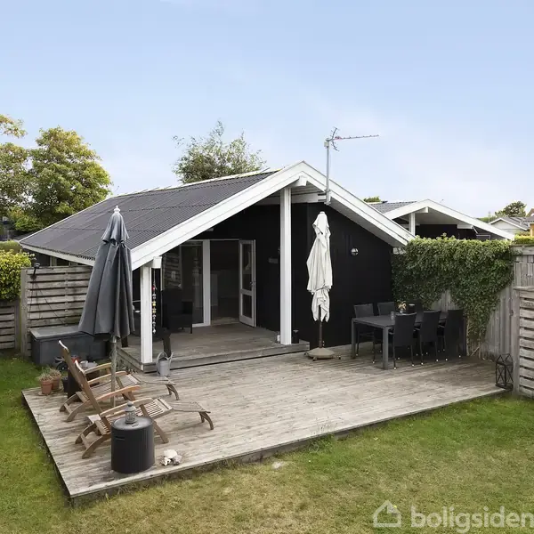 Et sort træsommerhus med en stor træterrasse, hvor der står havemøbler og en parasol. Haven er grøn og omgivet af hegn og buske under en klar blå himmel.