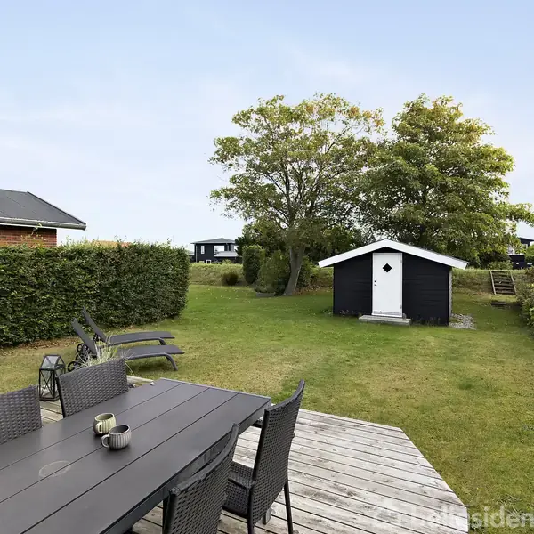 Sort skur står stille i grøn have med træer og buske. Terrasse i forgrunden med bord, stole og liggestole.