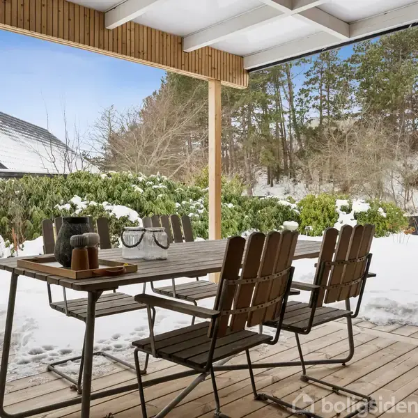 Udendørs træterrasse med spisebord og stole dækket af sne. Bordet har dekorationer. Omgivet af snedækkede buske og et hus med mørkt tag i baggrunden.