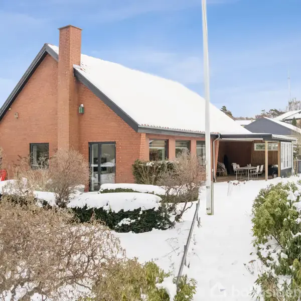 Et rødt murstenshus dækket af sne står blandt buske og træer i et snedækket landskab. Huset har store vinduer og en veranda med havemøbler. Himlen er blå.