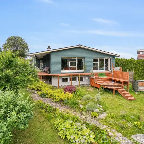 Et moderne hus har to store vinduer og en træterrasse. Det ligger i en velplejet have med grønne buske og græsplæne. Terrasse har trætrapper.