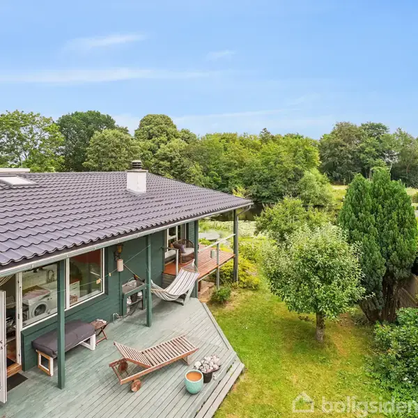 Hus med terrasse og solsenge ligger ved en frodig have med træer og en dam i baggrunden. Skyfri blå himmel skaber en rolig atmosfære.