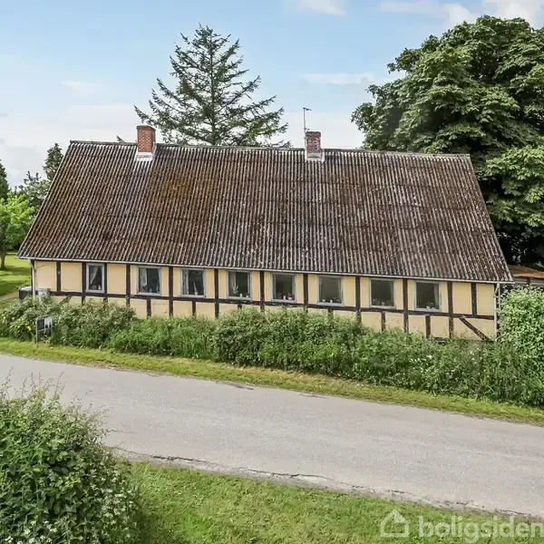 Et langstrakt bindingsværkshus med sort tag stående langs en vej. Huset er omgivet af grønne hække og træer i landskabelig omgivelser. Der er en trætopt i baggrunden.