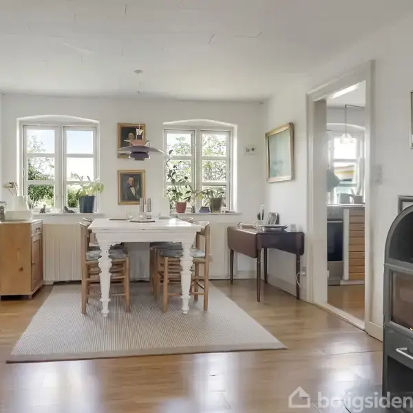 En spisestue har et spisebord med stole omkring. I baggrunden er der store vinduer, og på højre side er der en tændt brændeovn. Indretningen er lys og indbydende.